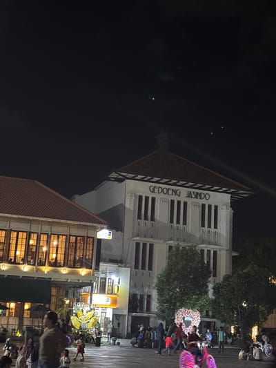 The image depicts a night scene in Fatahillah Square, a public square in the Old Town of Jakarta (Kota Tua), West Jakarta, Indonesia. In the background, a large white building with the prominent inscription "GEDOENG JASINDO" is illuminated. Adjacent to it, to the left, is another building with a reddish-brown tiled roof and windows with warm interior lighting. The square itself is paved and populated by a moderate number of people. Several individuals are visible walking, standing, and sitting. In the mid-ground, there is a large, ornate floral decoration, possibly a wreath or display, with a heart shape within it. To the right of the floral display, a person is riding a brightly lit pink bicycle. Various smaller stalls or shops with illuminated signs are also present, including one with the "SPARK" logo. The sky above is dark, with faint celestial lights visible. The overall atmosphere suggests an evening gathering or event in a historical urban setting.