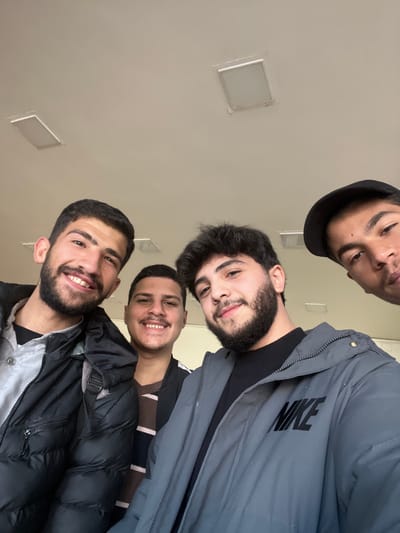 This image captures a selfie of four young men indoors in the city of Rukn ad-Din Municipality, Syria. The scene is illuminated by artificial light from two rectangular flush-mounted ceiling fixtures on a light-colored ceiling, which occupies the upper half of the frame.
From left to right:
1. A young male with dark hair and a short beard is smiling. He wears a black puffer jacket over a light grey collared shirt. A black strap is visible on his left shoulder.
2. Slightly behind the first individual, another young male with short dark hair and a mustache is smiling. He is wearing a dark, possibly striped, top.
3. In the foreground, a young male with dark wavy hair and a full beard is smiling. He wears a grey puffer jacket with the word "NIKE" printed in black on the right chest area, over a black ribbed shirt.
4. Partially visible on the far right, a fourth young male is present, with only the upper portion of his face and a black baseball cap visible. He is looking directly at the camera.
All individuals appear to be in their late teens or early twenties and are positioned closely together, facing the camera.