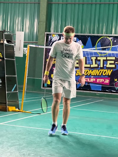 A young man is playing badminton indoors. He is wearing a white t-shirt with "Nike Track Club" printed on it, light gray shorts, a light green headband, and blue badminton shoes. He is holding a blue badminton racket and is in the middle of a game. The court is green with white lines. In the background, there is a banner that reads "ELITE BADMINTON CUP EP.2". The walls of the facility are green. The image appears to be taken at a badminton court in Ban Rang Rak, Thailand.