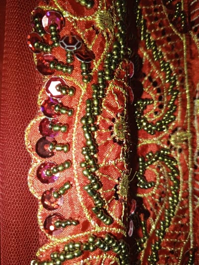 This close-up image depicts a section of an elaborately decorated red textile, located in Keo Kouang, Laos. The scene focuses on intricate embellishments across a rich red fabric background. To the left, a finer, sheer red mesh-like fabric is visible. Adjacent to this, a scalloped border crafted from opaque red fabric is adorned with numerous reflective, iridescent red sequins of both circular and hexagonal shapes. Each sequin is secured with small clusters of metallic gold-colored beads. To the right of a central vertical fold or seam, a denser opaque red fabric is extensively embroidered with gold-colored thread forming complex curvilinear patterns, possibly floral or paisley motifs. Lines and clusters of additional gold-colored beads are integrated into these embroidered designs, highlighting and accentuating the patterns. The overall impression is one of detailed craftsmanship, with varying textures and reflective elements catching the light. No people or active interactions are present; the image is a static portrayal of the textile's surface.