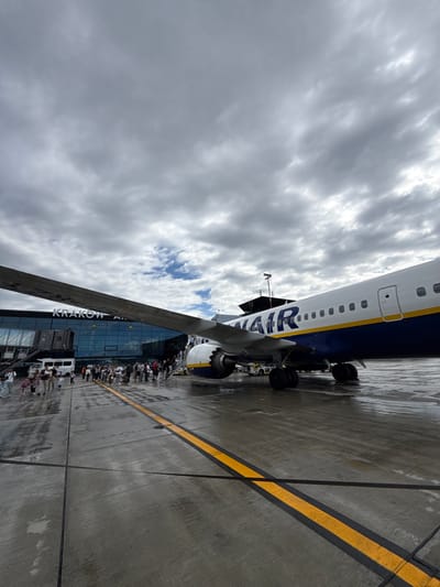 The image depicts Krakow John Paul II International Airport in Balice, Poland, on a cloudy day. A Ryanair airplane, predominantly white with a blue and yellow livery, is parked on the tarmac. The wing of the aircraft extends prominently into the foreground, partially obscuring the modern airport terminal building, which features a glass facade and prominently displays the text "KRAKOW AIR." A jet bridge connects to the terminal. Several people, including adults and children, are seen disembarking the aircraft via stairs or walking towards it. Some are carrying luggage. Service vehicles, including a tug, are visible on the wet tarmac, indicated by reflections and damp patches on the concrete. Yellow and black lines mark the runway. The overall scene captures the activity of passengers boarding or disembarking an aircraft at an airport.