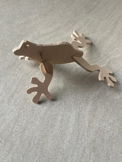 The image shows a light brown, wooden, three-dimensional cut-out of a stylized gecko or lizard. It is positioned on a light grey, speckled surface, likely a countertop or textured wall. The gecko is oriented vertically in the frame, with its head pointing downwards towards the bottom left corner. Its body is flat, and its limbs are articulated and spread out, suggesting it is either climbing or in a resting position. The gecko has four limbs, each ending in five finger-like digits. A small circular hole is visible near the head, possibly for hanging or as part of the design. The lighting casts subtle shadows, indicating the light source is from the upper right. The overall scene is simple and focused on the object, with no other prominent subjects or background details. There is no visible text in the image. The location context is Jelgava, Latvia, but there are no specific visual cues in the image to confirm this.