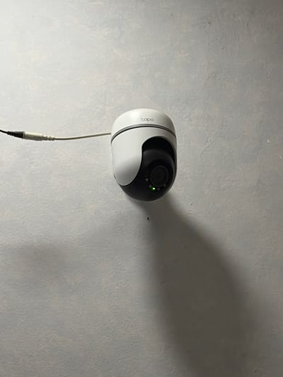 A security camera is mounted on a textured, light gray wall. The camera is white with a black lens housing. A small green light is visible on the front of the camera, indicating it is operational. A white cable with a black connector extends downwards from the camera, suggesting it is powered or connected to a recording device. The camera is positioned in a way that casts a large shadow on the wall to its right, indicating a light source to the left of the frame, likely artificial. The scene is indoors, with the wall texture suggesting an indoor environment such as a room or hallway. There is no visible indication of the time of day or weather conditions. The word "tapo" is visible in white text on the side of the camera. No people are present in the image.