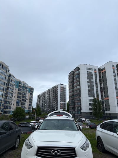 The image captures a street scene in Tokkari, Russia, under an overcast sky. Several apartment buildings with distinct architectural designs line the background. The lower buildings feature light-colored facades with blue and brown balcony accents. Higher buildings have white and grey facades with smaller, repetitive window arrangements. A row of parked cars occupies the foreground, with a white Infiniti QX60 prominently positioned on the right. A black sedan is visible on the lower right, and other cars are parked along the street. The scene suggests a residential area with dense housing.