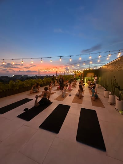 The image depicts a yoga class taking place on a rooftop at sunset in Tibubeneng, Indonesia. Approximately 20-30 people are participating in the class, positioned on mats laid out on a tiled terrace. Most of the participants are performing yoga poses. The sky transitions from blue to orange and pink hues as the sun sets. String lights are strung along the edge of the rooftop, adding to the ambiance. Vegetation is visible below the terrace, suggesting an elevated location. Potted plants are placed along the perimeter.