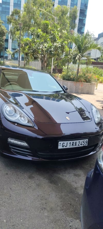 A dark-colored Porsche Panamera is parked on an asphalt surface in an urban setting. The car's license plate reads "GJ 1 RA 2459". In the background, there is a modern office building with glass facades, and there is greenery including trees and bushes in planters, and a fence. Another vehicle, partially visible on the right edge of the frame, appears to be dark blue. Based on the license plate (GJ), the car is registered in Gujarat, India. The setting, a modern office park with palm trees and manicured landscaping, suggests a corporate area within a major Indian city, possibly Mumbai.