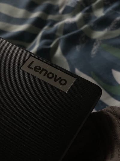 The scene is located in Saverne, France. A black Lenovo electronic device occupies the mid-ground, observed from an oblique, close-up perspective. The device's visible surface exhibits a fine, repeating grid texture. Centrally located on this surface is a rectangular metallic plate with the word "Lenovo" embossed in black capital letters. The device is positioned against a soft, wrinkled textile background patterned with horizontal stripes in varying shades of dark blue and lighter blue or white. A portion of a dark grey or brown fabric, likely part of a garment, is visible along the lower right edge of the frame. No individuals or active interactions are depicted.