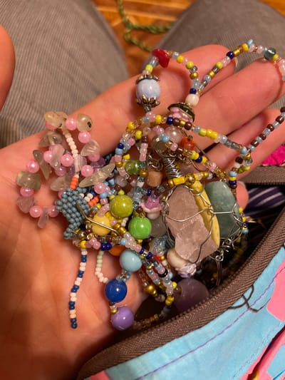 A close-up, over-the-shoulder shot shows a person's hand holding a jumble of colorful, handmade beaded jewelry. The jewelry consists of a variety of beads, including small seed beads, larger opaque beads in shades of pink, blue, purple, and green, and some larger translucent stones, possibly quartz or agate, wrapped in wire. The beads are strung together in necklaces, bracelets, or possibly part of a larger craft project. The background is out of focus, but appears to be a textured surface, possibly fabric, in shades of grey. The jewelry is spilling out of a bag with a bright blue and pink patterned fabric. The image is well-lit, highlighting the textures and colors of the beads. There is no discernible location within Sevastopol, Ukraine, as the image focuses entirely on the hand and the jewelry.