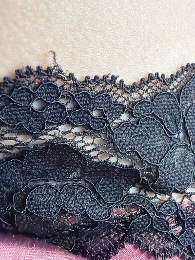 This is a close-up, abstract shot of black lace fabric. The lace has a floral or leaf-like pattern with intricate openwork. Part of the lace is layered over a pale, textured surface, possibly skin or a light-colored fabric. The other part of the lace is against a darker, possibly pink or purple satin-like fabric. There are no people, activities, or specific location cues visible. The image focuses solely on the texture and detail of the lace.