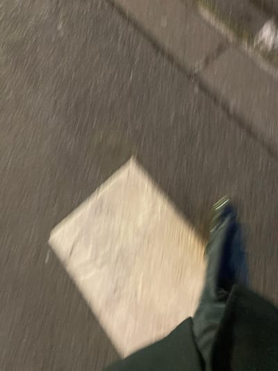A blurred image shows a section of a sidewalk, composed of dark grey stone blocks. A roughly square patch of a lighter, cream-colored material is set into the sidewalk. The edge of a dark shoe and dark green fabric are visible in the upper right corner, suggesting a person is standing nearby. The blurry quality of the image gives a sense of motion.
