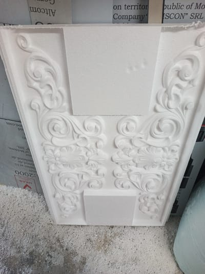 This media file displays a close-up, slightly angled view of a white, intricately carved foam object, likely a decorative molding or architectural element. The carving features elaborate swirling scrollwork and floral motifs, creating a sense of classical ornamentation. The object is positioned on a surface covered in fine white debris, suggesting it is a workshop or manufacturing environment where foam is being cut or shaped.

In the background, visible behind the foam object, are sheets of paper with printed text. Some of the text is partially obscured or out of focus, but fragments of words and company names can be deciphered. Notably, the words "Altcom," "on territory," "Company," "public of Mo," and "ISCON" SRL are visible. Further down, a label with "UKAS :2000" and "nbH" is present, along with what appears to be a company website or address fragment ending in ".com".

The lighting appears to be artificial, casting no distinct shadows that would indicate a specific time of day. The overall impression is one of craftsmanship and production in a professional setting. The location context provided indicates Durlești, Moldova.