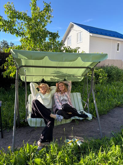 Two women are relaxing on a green patio swing in a backyard. The woman closer to the viewer is wearing a light-colored sweatshirt and black pants. The woman behind her is wearing a pink and white plaid shirt. Both women are lying down on the swing. The swing has a green canopy and is set on a gravel path surrounded by lush green grass and trees. A white house with a blue roof is visible in the background, along with a clear blue sky. The sunlight suggests it is a bright, sunny day, likely late afternoon given the long shadows. The overall scene conveys a sense of relaxation and enjoyment of the outdoors in a residential setting.