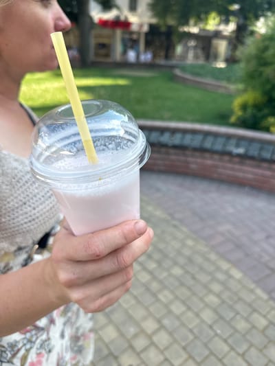 A close-up, low-angle shot shows a person holding a clear plastic cup filled with a light pink liquid, likely a milkshake or smoothie. A yellow straw extends from the cup. The person's hand and arm are visible, with a glimpse of their face and shoulder in the lower left corner. They are wearing a light-colored, knitted top over a floral-patterned garment. The background is blurred, showing greenery and a brick wall, suggesting an outdoor setting, possibly a park or garden. The lighting indicates it is daytime. The image appears to have been taken in Haskovo, Bulgaria.