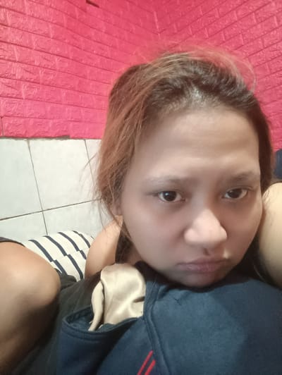 A woman, with her head resting on what appears to be a blue jacket or blanket, is looking directly at the camera. She has light brown hair and is wearing a dark blue jacket with red stripes on the sleeve. The background consists of a textured pink wall and white tiled wall. The scene is likely indoors, possibly in a bedroom or living room in Depok, Indonesia.