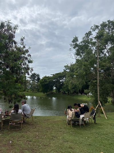 People sit in chairs on green grass beside a pond. Lush trees surround the water, with a cloudy sky overhead. Some people are gathered closer to the water's edge, while others are further back. The chairs are simple folding chairs. The setting has a peaceful, outdoor ambiance.