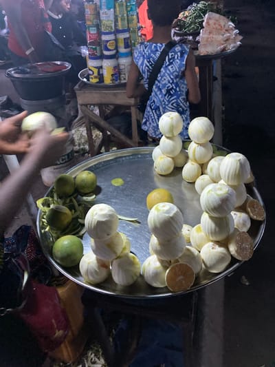 The scene depicts a market activity in Ura Nta Umuarandu, Nigeria, under low light conditions, likely evening or night. In the immediate foreground, a pair of dark-skinned hands is actively peeling a light-colored, spherical fruit over a large, round, metallic tray. The tray contains numerous other fruits, including multiple light-colored, peeled spherical fruits stacked in small pyramids (three visible stacks of approximately three to four fruits each), individual peeled fruits, several unpeeled green citrus fruits, and scattered discarded peels. Two peeled fruits on the tray are cut in half, revealing a pale orange interior.
In the midground, a person, possibly a child or young adult, stands with their back mostly to the camera, wearing a blue and white patterned sleeveless top with a dark strap across the shoulder. This individual faces a stall displaying various consumer goods, including stacks of canned vegetables (e.g., peas) and jars of mayonnaise on a wooden stand. To the right of this stall, another display holds green leafy vegetables and items packaged in clear plastic bags on a tray.
In the left background, a person in a red garment is partially visible, facing right. Above this individual, another person's head and shoulders are dimly seen. A black basin or pot rests on a surface to the left of the canned goods display. The ground surface appears unpaved, with debris visible.
