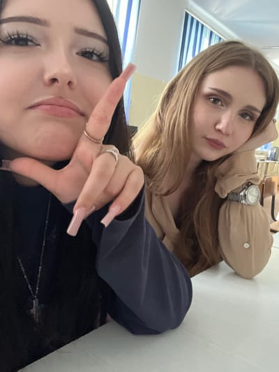 Two young women are seated at a white table, likely in a classroom or common area. The woman on the left is closer to the camera, her face framed by dark hair. She is looking directly at the camera with a slightly pouty expression, her right hand raised, fingers extended with long, light pink acrylic nails. She wears a dark top and a silver necklace with a cross pendant. On her fingers, she has two rings, one plain gold band and a more elaborate silver ring with stones. The woman on the right has long, light brown hair and is looking towards the left. She rests her head on her left hand, which is propped up by her elbow. She is wearing a light brown long-sleeved shirt with a button near the cuff, and a silver wristwatch with a circular face. The background shows a window with blue and white striped blinds, and the corner of a desk or computer monitor is visible on the far right. The scene is well-lit, suggesting it is daytime. No specific landmarks in Sterlitamak, Russia are identifiable from the image.