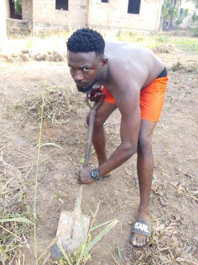 A man with dark hair and a beard is shown outdoors, shirtless and wearing bright orange shorts, digging with a shovel. He looks directly at the camera with a stern expression. He wears a watch on his left wrist and black sandals with "KARL" printed on them. The ground is dry and covered with grass and weeds. In the background is a building with visible windows. The overall impression is one of physical labor.