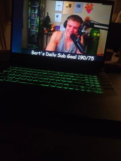 This is a close-up, eye-level, slightly angled indoor shot of a laptop screen displaying a live stream. The laptop has a green-backlit keyboard visible in the foreground, mostly in shadow. The screen shows a man with headphones and a microphone, smiling and looking towards the camera. Behind him, a green cartoon character resembling Rick Sanchez from Rick and Morty is visible, along with various framed awards on the wall. Text overlays on the screen read "Bart's Daily Sub Goal 190/75". The background is dark, indicating the room is dimly lit. There is no discernible information about the city of Versailles, France.