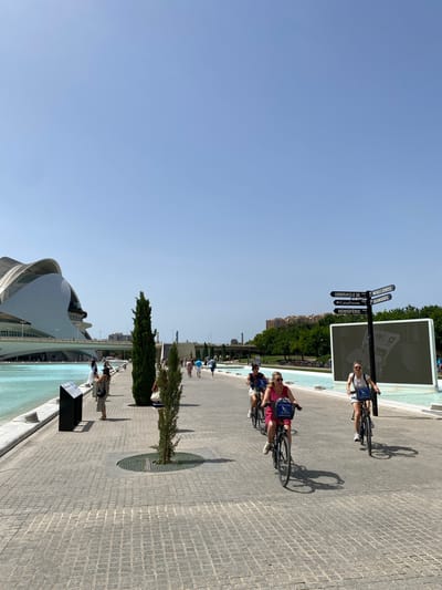 The image depicts a sunny day in the City of Arts and Sciences in Valencia, Spain. The foreground shows a paved walkway where three women are riding bicycles. The woman in the center wears a pink dress, the one on the left wears a black top and shorts, and the woman on the right wears a white top and shorts. All three have baskets on their bikes. In the middle ground, there are two tall, slender trees and a signpost with directions to "UMBRACLE," "CAIXAFORUM," "HEMISFERIC," "MUSEU DE LES CIENCIES," and "OCEANOGRAFIC." Behind the signpost is a large, flat screen displaying an image. To the left, a modern architectural building with white, curved structures and a bridge is visible, adjacent to a bright turquoise pool of water. Several other pedestrians are walking in the background, and a line of trees stretches towards the horizon. The sky is clear and blue.