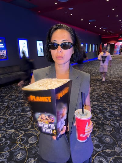 This media file features a medium shot of a woman wearing large, dark sunglasses, holding a large popcorn container and a red Coca-Cola cup with a straw. She is seated in what appears to be a cinema or entertainment venue. The background shows a dark, carpeted floor with a circular pattern, and the characteristic dark red walls and glowing illuminated signs commonly found in cinemas. The woman is looking slightly upward and to the side, with a neutral expression. The popcorn container has bold orange text that reads "PLANET" and some Hebrew text along with cartoon characters. The Coca-Cola cup is iconic with its red color and white logo. The overall scene suggests a casual outing to watch a movie. The lighting is dim, typical of a pre-movie or intermission setting in a cinema. The woman is dressed in a grey blazer, over dark clothing. The image is taken from a slightly low angle.
