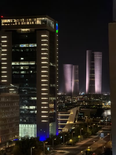 The photograph captures a nighttime view of Lusail, Qatar, showcasing a modern urban landscape dominated by illuminated skyscrapers and architectural structures. In the foreground and left of the frame, a tall, rectangular building features a facade lined with horizontal, illuminated strips and numerous windows, some of which reveal lit interior spaces. To the right of this building, a series of sleek, cylindrical towers rise, their surfaces illuminated with a textured, purplish light, creating a striking visual effect.
Below the towers, the city unfolds with a network of roads, streetlights, and lower-rise buildings. The streetlights cast warm yellow glows on the pavement and sidewalks, while trees and landscaping add touches of green to the urban scene. One notable building in the mid-ground has a distinctive, angled roof design and is brightly lit, highlighting its modern architecture.
The sky is dark, emphasizing the artificial illumination of the city. The overall impression is one of a vibrant, technologically advanced city at night. There are no visible people or discernable human activity in the captured scene. The image focuses on the architectural elements and the ambient lighting that defines the urban environment of Lusail.