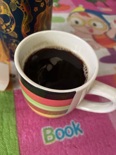A high-angle shot depicts a colorful coffee mug filled with dark liquid, likely coffee, resting on a multi-colored surface. The mug is mostly white, with a vertical series of stripes in shades of pink, red, green, and yellow. A white handle is attached to the mug. A portion of a decorative object with a dark blue base and gold embellishments is visible at the bottom left. The background surface features cartoon-like figures in bright colors, including pink, green, and yellow. There is a text "book" printed in large blue letters on the pink surface. There is no verifiable information linking this image to the city of Hijo, Philippines.