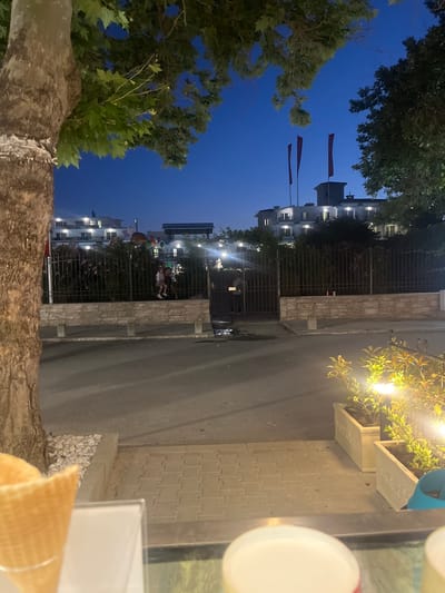 The image shows a street scene at dusk in Sozopol, Bulgaria, with a focus on a large tree trunk in the foreground on the left. Beyond the tree, a paved street is visible, leading towards a fence that separates the street from a complex of buildings. The buildings appear to be hotels or apartments, with windows lit from within. Three red flags are flying from the top of one of the buildings. Two individuals, possibly children, are standing near the fence, looking towards the camera or away from it. The sky is a deep blue, indicating twilight. In the foreground, an ice cream cone is partially visible, suggesting the presence of an ice cream shop or stand. Planters with illuminated foliage are also present on the right side of the image. The overall atmosphere is calm and serene, with the lighting suggesting evening.