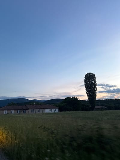 The image depicts a rural landscape near Slokoshtitsa, Bulgaria, at dusk. The sky is a gradient of clear blue at the top transitioning to lighter hues near the horizon, with some scattered clouds. In the middle ground, a long, single-story house with a tiled roof is visible, featuring multiple windows and air conditioning units. Adjacent to the house is a field of tall, golden-brown grass. To the right of the house stands a very tall, slender tree. Behind the house and grass, a line of dark green, undulating hills forms the background, with dense foliage and some scattered structures. The overall impression is one of a quiet, rural evening.