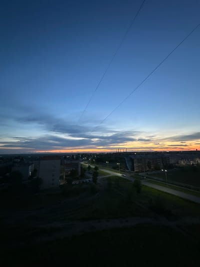The image depicts a twilight view of a city, possibly Yuzhnouralsk, Russia, looking eastward. The sky displays a gradient of colors, transitioning from deep blue overhead to hues of orange and yellow near the horizon, indicating either sunrise or sunset. Clouds are visible, scattered across the sky and reflecting the warm light from below. In the foreground, there is a dark, grassy slope. Below the slope, a street is visible with streetlights illuminating the roadway and the side of the road. Several multi-story residential buildings are situated along the street, with some windows appearing lit. Cars are parked near the buildings and a single car is driving on the road. Power lines are visible stretching diagonally across the sky from the upper left to the upper right. The overall scene suggests a quiet evening or early morning in a residential area.
