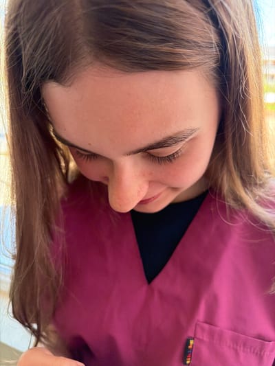 A close-up, low-angle shot depicts a young woman with fair skin and light brown hair. Her eyes are closed, and she appears to be resting. She is wearing a pink top with a black collar. The location, Panagyurishte, Bulgaria, is not discernible from the image.