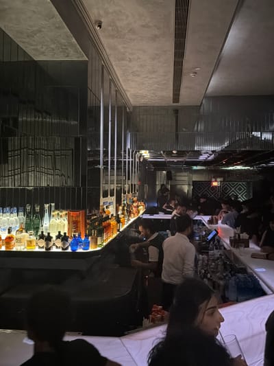 The image captures an indoor scene, likely a bar or lounge, in New Delhi, India. A long, well-stocked bar dominates the foreground, lined with an extensive collection of liquor bottles. Several bartenders are visible behind the bar, some appearing to be serving customers. Patrons are seated at the bar or standing nearby, engaged in conversations or observing the scene. The interior design features dark, reflective surfaces, modern lighting, and a metallic grillework in the background, suggesting a contemporary and upscale establishment. The overall atmosphere appears to be lively and social, characteristic of a nightlife venue.
