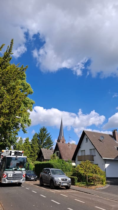 A street scene in Cologne, Germany, under a blue sky with scattered white clouds. The image features a silver Kia Sorento parked on the right side of the street, facing left. To its left, a black Mercedes-Benz is parked. Further left, a silver utility truck is partially visible. In the background, several residential houses with brown tiled roofs are situated behind lush green hedges. A church steeple with a dark spire rises above the houses. A large tree with green foliage is prominent on the left side of the frame. The street is asphalt with white lane markings.