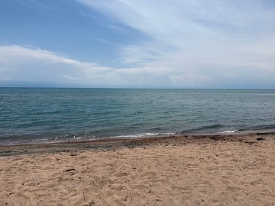 A wide-angle shot captures a tranquil beach scene in Kara-Oy, Kyrgyzstan, under a partly cloudy sky. The foreground is dominated by a sandy beach, with footprints and scattered debris like twigs and small stones visible. The sand is a light tan color, showing ripples and textures from natural elements.

Beyond the beach lies the calm, blue water of a lake, its surface reflecting the sky and shimmering with sunlight. Gentle waves lap at the shore, creating a foamy white line where they meet the sand. The water appears clear and inviting.

In the distance, the horizon is marked by a faint line where the water meets the sky, suggesting a vast expanse. The sky above is a bright blue, streaked with wispy white clouds. These clouds are soft and diffuse, with some appearing more solid and clustered, hinting at a pleasant, possibly warm day. The overall impression is one of peaceful natural beauty, characteristic of a lakeside resort area like Kara-Oy. There are no people or man-made structures visible in this specific frame.