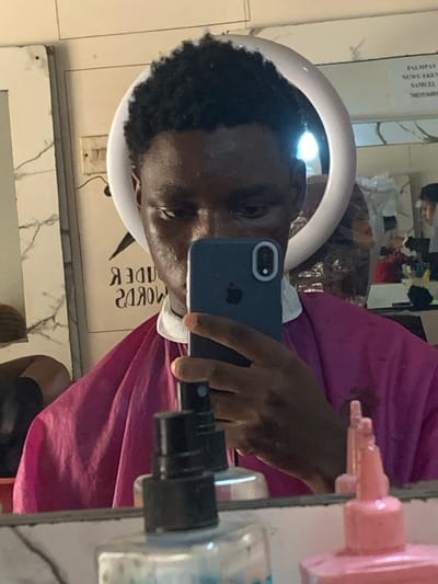 The scene depicts a male individual taking a mirror selfie in what appears to be a barbershop or salon in Eziobodo, Nigeria. The individual, who has dark skin and short, dark, curly hair, is partially obscured by a dark blue smartphone held in his right hand. He is wearing a magenta-colored barber's cape with a white collar.

Reflected in the mirror are several objects and details:
A white circular ring light is positioned behind the individual's head, indicating artificial illumination.
To the left, reversed text from a sign reads "ЯЗ ADWARDS," likely "AWARDS ЗЯ" or similar in its original orientation. A faint, dark silhouette, possibly another person or a mannequin head, is also visible.
To the right, another reflected sign displays text including "PALMPAY," "NGWU EK[E/O]N," "SAMUEL," and a partial phone number "708353680[1/4]," suggesting business or contact information.
On a counter below the mirror, various bottles are present, including a black spray bottle with a blue nozzle, two pink-capped squeeze bottles, and a clear bottle containing a blue liquid. The background wall features white or light-colored tiling, with a marbled pattern visible on the left side.