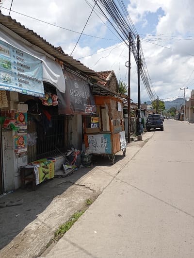 The scene is a street in Katapang, Indonesia, located at latitude -6.998 and longitude 107.552. The street is paved and lined with buildings on both sides. There are numerous power lines running overhead. On the left side of the street, a small wooden cart with a sign that says "MAGNUM KRITE" is parked near a building with a sign advertising snacks, cookies, frozen food, soap, household goods, and cosmetics. In front of the cart, there is a pile of discarded items, including a black bucket, a green bucket, a white bag, and a green bag. There is also a wooden bench with a white and yellow bag resting on it. On the right side of the street, a black car is parked on the shoulder of the road. The street is empty, except for the car. The sky is partly cloudy. The buildings are painted in muted colors and appear to be in disrepair. The overall atmosphere is quiet and peaceful.