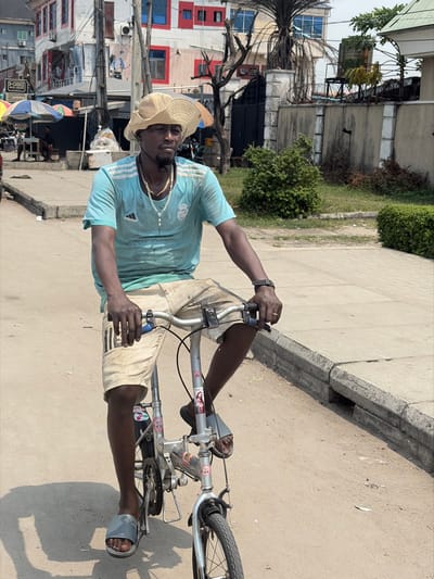A dark-skinned adult male is shown riding a silver-colored folding bicycle on an unpaved road in Jakande, Nigeria. He wears a straw hat, a light blue short-sleeved Adidas Real Madrid t-shirt, light-colored (possibly denim) shorts, and grey slide sandals. His face features a short dark beard, and he has black earphones/earbuds around his neck. He wears a gold-colored chain necklace, a white beaded rosary-style necklace, and a dark wristwatch on his left wrist. His hands grip the bicycle's handlebars, and his gaze is directed forward with a neutral expression. A sticker depicting "Jesus" is visible on the bicycle's frame. To the right, a concrete curb and sidewalk border the road. The background reveals an urban environment with multi-story buildings, including one with prominent red and white sections and blue-framed windows, and another with a dark metal gate. Multiple utility poles with numerous wires are present, alongside sparse vegetation like bushes and a palm tree. On the far left, colorful market umbrellas and a white container are visible, indicating a commercial area. The scene is brightly lit by daylight, casting shadows.