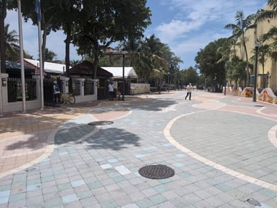 This media file captures a bright, sunny day in a pedestrian walkway in Miami, USA. The scene features a colorful paved pathway with intricate geometric patterns and alternating shades of grey, blue, and pinkish-tan bricks. Several manhole covers are visible on the ground.
In the background, a yellow bicycle leans against a black fence, next to a person wearing a white hat and blue backpack. Further down the path, another person is walking away from the camera, wearing a light-colored short-sleeved shirt and dark pants, with a light-colored cap. A white chicken is also present on the walkway.
The walkway is lined with trees, including palm trees, and architectural structures that appear to be buildings or shaded areas with awnings. A light-colored tent is set up on the left side of the image, and a yellow building with colorful decorative elements is visible on the right. Streetlights are spaced along the pathway. The sky is blue with scattered white clouds, suggesting it is daytime, likely mid-morning or afternoon. The overall atmosphere appears to be calm and leisurely, indicative of a public space designed for walking and enjoying the outdoors.