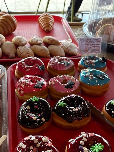 This image captures a close-up view of a bakery display in Tsovagyugh, Armenia, featuring a variety of baked goods arranged on red trays. In the foreground and mid-ground, multiple rows of ring-shaped doughnuts are presented on a lower red tray. The doughnuts feature various glazes: bright red, light pink, light blue, dark brown (chocolate), and dark red/maroon. Each doughnut is adorned with multi-colored sprinkles; some have additional decorations such as small black butterfly shapes (on red, pink, and blue doughnuts), green star shapes (on dark brown and dark red doughnuts), and small white or pink flower sprinkles. A silver serving tong is visible on the right, positioned next to a dark brown doughnut.
On an upper, larger red tray, two golden-brown croissants are displayed, one prominent on the left and another further back on the right. Below the croissants, several smaller, oblong-shaped pastries are visible. Some of these smaller pastries are plain, light brown, while others have a light-colored glaze with a red zig-zag drizzle pattern. A clear plastic pricing label is affixed to the upper red tray on the right side. The label contains text in Armenian and English, clearly stating "BERLIN CHOCOLATE BAR" and "380դր" (380 Armenian Dram), along with a numerical code "9444". The background is brightly lit, showing blurred architectural elements that could be window frames or railings, and the lower legs and feet of a person wearing dark pants and light-colored shoes are partially visible on the right, indicating an indoor commercial setting such as a bakery or market.