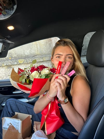 A young Caucasian female with blonde hair is seated in the front passenger seat of a dark-colored vehicle. She wears a black sleeveless top and blue jeans, with a watch featuring a brown leather strap on her right wrist and multiple thin bracelets on her left. She is smiling directly at the camera while holding a bouquet of red and white roses, wrapped in brown paper and red fabric/ribbon. In her right hand, she also displays three "rhode" brand lip tints: one red, one light pink, and one darker pink. On the vehicle's floor in front of her, a partially opened cardboard box with internal brown paper packaging material and a white plastic bag are visible. The car interior features a black headliner and black leather seating. Through the passenger window, a textured concrete or stone wall and green foliage are visible under bright daylight, indicative of an outdoor location in Cidade da Maia, Portugal.
