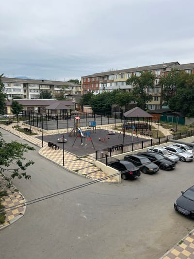 This is an aerial, eye-level, medium shot capturing a residential courtyard in the city of Buynaksk, Russia, on an overcast day. The scene is dominated by apartment buildings, a playground, and a parking lot filled with cars.
The apartment buildings are typical of Soviet-era architecture, with multiple stories, uniform windows, and balconies. They appear to be in varying states of repair, with some showing signs of age and others appearing more recently renovated with new siding or paint. The buildings are arranged in a U-shape around the courtyard, creating a sense of enclosure.
In the center of the courtyard is a fenced-in playground with swings, a slide, and a merry-go-round. The playground is surrounded by a rubberized safety surface. Adjacent to the playground is a covered seating area with benches.
A paved road curves through the courtyard, with cars parked along its edges. The cars are a mix of makes and models, and most appear to be sedans. The road is lined with sidewalks and a checkerboard pattern of yellow and beige tiles.
The environment is somewhat urban and densely populated, as evidenced by the number of apartment buildings and cars. The overcast sky suggests a cooler climate, and the presence of trees indicates a temperate region. There are no people visible in the image.