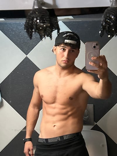 A male individual is depicted in a mirror selfie, shirtless, revealing an athletic physique with visible abdominal muscles. He has short, curly dark hair and is wearing a black baseball cap with white reversed text on the front. On his left wrist is a black digital watch. He is also wearing black athletic shorts or pants with a visible "PUMA" waistband, which appears reversed in the reflection. The subject holds a light purple smartphone with his right hand, angled to capture the image in the mirror; the phone exhibits a multi-lens camera array and a circular magnetic attachment on its back. His expression is neutral, and his gaze is directed towards the phone's reflection. The background consists of a wall tiled with black and white squares arranged in a diamond pattern. To the lower right, part of a white ceramic fixture, possibly a toilet or urinal, is visible, with a silver flush mechanism on the wall above it. Reflected in the upper background are two dark, textured, possibly decorative objects that appear to be hanging. The specific geographic location in Sarouja Municipality, Syria, cannot be determined from the visual information provided.