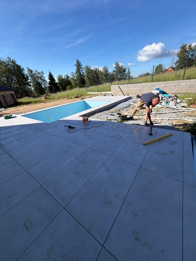 The image depicts an outdoor construction scene near a swimming pool in Horní Planá, Czechia. A man wearing a dark t-shirt and khaki shorts is bent over, working on the tiled patio. Tools, including a spirit level and what appears to be a trowel, are visible near him. The patio is partially tiled with large, light-colored square tiles, some of which show reddish-brown smudges. An unfinished swimming pool with blue lining is to the left of the patio. In the background, there is a retaining wall constructed from concrete blocks, piles of building materials, and a chain-link fence bordering a grassy area with trees. The sky is blue with scattered white clouds, and the sun is shining, casting shadows.