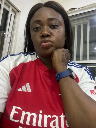 A woman is pictured indoors, her face angled towards the camera. She has dark hair, dark eyes, and a nose piercing. She rests her head on her hand, which is adorned with a tattoo. She wears a red and white soccer jersey with "Emirates" printed on it. A blue smartwatch is on her wrist. A window frame is visible in the background. The location in Kubwa, Nigeria cannot be determined from the image.