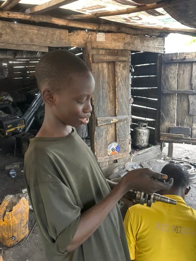 In the city of Badagry, Nigeria, a young boy in a green t-shirt stands in what appears to be a workshop, holding a metal crankshaft. His gaze is directed downwards towards the crankshaft, and his mouth is slightly open as if he is speaking or explaining something. Behind him, another person wearing a yellow t-shirt is visible from the back. The workshop environment is rustic, with wooden structures forming the walls and ceiling. Various tools and equipment, including what looks like a generator and a gas cylinder, are scattered around the dusty ground. The scene suggests a working environment, possibly a mechanic's shop, where the young boy might be assisting or learning.