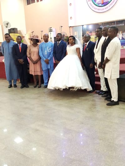 A wedding photo captures a group of individuals, likely family and friends, flanking a bride in a voluminous white gown. The diverse group includes men in suits of varying colors and women in dresses and hats. They stand on a glossy floor in what appears to be a church, suggested by the elevated stage and a sign with religious text in the background. The lighting is bright and natural.