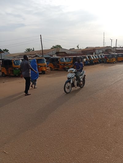 A scene unfolds on a paved road in Nassarawa, Nigeria. A row of motorized rickshaws, commonly known as "Keke Napeps," are parked on the side of the road. A man in a grey shirt and black pants walks alongside a figure cloaked in a blue hijab. A man on a white and blue motorcycle drives towards the viewer. In the background, buildings with corrugated metal roofs are visible, along with a communications tower. The sky is overcast.