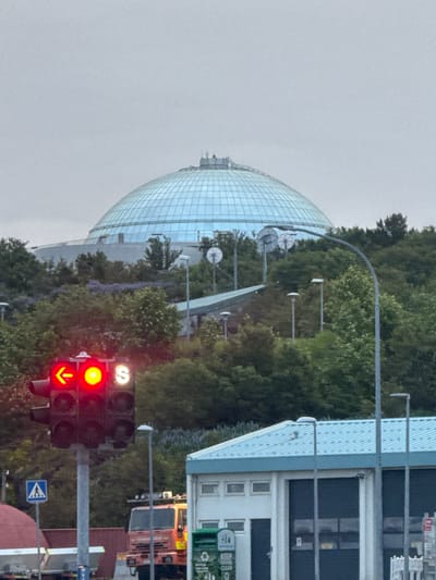 This image captures an exterior view of the Perlan museum and observation deck in Reykjavik, Iceland. The iconic glass dome, a prominent feature of the building, is visible in the upper portion of the frame, reflecting the overcast sky. The surrounding landscape is characterized by lush green trees and foliage, with a hint of purple flowering plants. In the foreground, a traffic light displays a red left arrow and a yellow light, indicating a restriction on turning left. Next to it, a white "S" symbol is illuminated, possibly signifying a bus stop or other public transportation indicator. To the right, a modern building with a light blue roof and large dark garage doors is partially visible. A red fire truck is parked in front of this building. A pedestrian crossing sign with a blue background and a white triangle containing a figure walking is also present. The overall atmosphere is somewhat muted due to the cloudy weather.