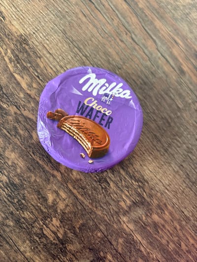 A single, round, Milka Choco Wafer snack, still fully encased in its purple foil wrapper, rests diagonally on a wooden surface. The wrapper displays the white "Milka" brand logo, accompanied by a small white cow icon, and the product name "Choco Wafer" in gold lettering. An illustrative image on the wrapper depicts a chocolate-covered wafer bar, broken to reveal its multiple light-colored wafer layers and brown chocolate filling, with small crumbs scattered around the depicted wafer. The underlying surface is composed of distressed wooden planks, featuring varied shades of brown, black, and tan, with visible diagonal grain patterns running from the top-left to the bottom-right. No people or other subjects are present, and no actions or interactions are depicted. The scene is stated to be located in the city of Ihtiman, Bulgaria.