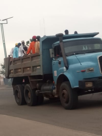 A light blue dump truck is carrying several men in its open bed. The men are wearing traditional clothing, including colorful shirts and hats. Some of the men are standing, while others are seated. The truck is driving on a paved road, with a hazy sky and some vegetation in the background. A solar-powered street light is visible on the left side of the image. The scene appears to be in Pantami, Nigeria.